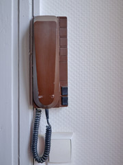 Vintage brown wall phone, hanging off a white textured wall.  Phone also has intercom bottoms to let people in front door