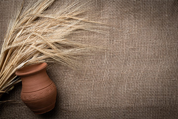 Yellow dry spikelets of wheat on a burlap with a clay pot. Place for text. Still life close up. Top view.