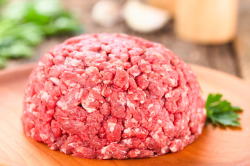 Fresh raw ground or minced beef meat on wooden plate, seasonings (salt, pepper), garlic and fresh...