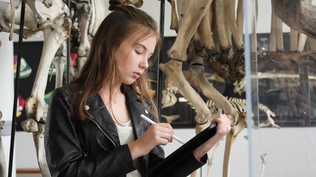 Teenager cute girl makes draw sketches in graphic tablet of animal skeletons in museum of nature as part of school task of biology