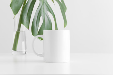 White mug mockup with a monstera leaf in a glass vase on a white table.