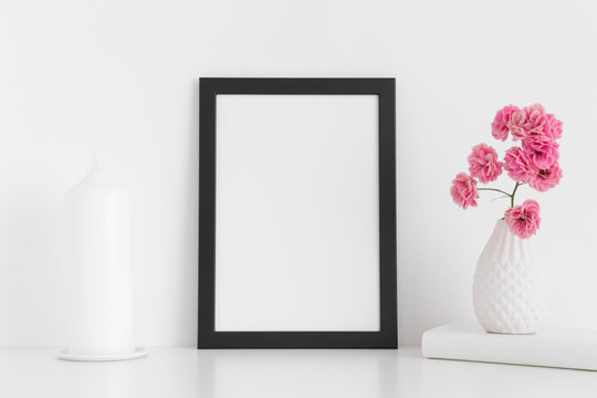 Black frame mockup with pink roses in a vase and candle on a white table.Portrait orientation.