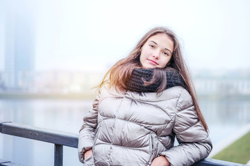 modern young girl teenager stands on the bridge leaning on the railing hiding her hands in the pockets of a warm jacket