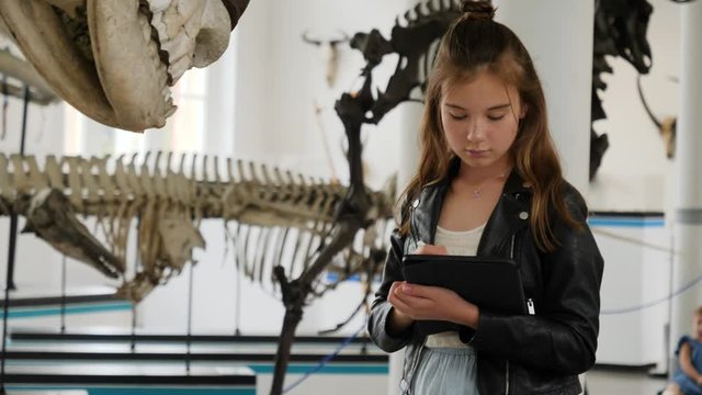 Teenager cute girl makes draw sketches in graphic tablet of animal skeletons in museum of nature as part of school task of biology