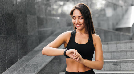 Young smilling fit woman looking at smart bracelet on background of gray marble stairs outdoor. Sports Gadgets