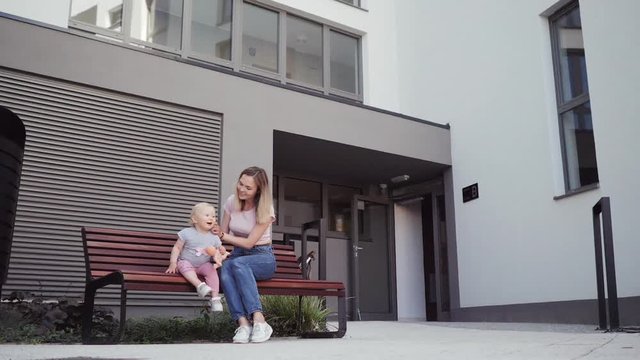 young mother walks with her little girl daughter around the house sitting on a bench talking
