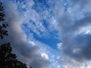 Landscape of beautiful blue sky with light thin fluffy white clouds and the gap between the thunderclouds with the sun illuminates part of the clouds and trees on the edges. 