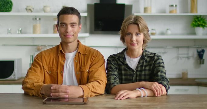 Portrait of young latin man and white woman, smiling at camera, sitting at table in kitchen, looking confidently in their future, happy, wearing casual clothes. 4K, shot on RED camera.