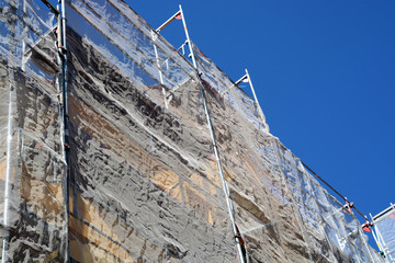 protection grid and scaffolding covered old house facade during renovation in front of azure sky,...