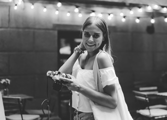 Cheerful blond woman in fashionable clothes holding retro camera in evening on the background of...