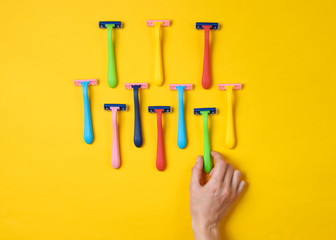 Female hand choosing green razor among a variety of colored epilator razors on yellow background. Top view. Minimalism beauty concept. Top view