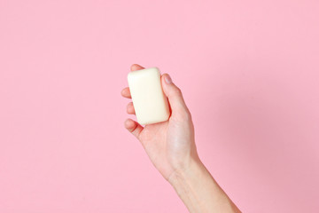 Female hand holding piece of soap against pink background. Top view