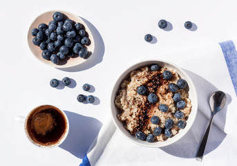 Oatmeal with blueberies and cocoa powder in a round bowl, plate full of blueberries and a cup of black coffee on a napkin. Top view.