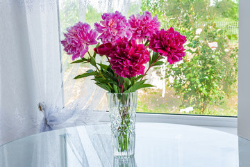 A large bouquet of pink flowers peonies standing in the room on the table in a crystal vase by the window