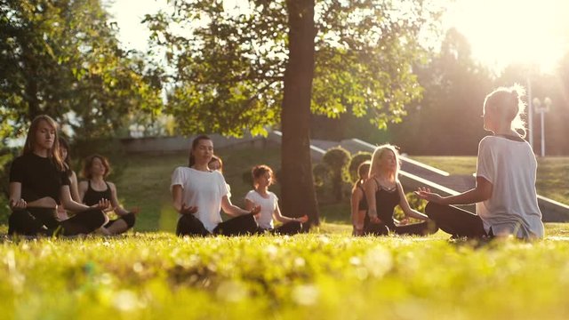 Group of young women meditate in park on summer sunny morning under guidance of coach in slow motion. Group of girl outdoors is sitting in lotus pose on green grass with their eyes closed