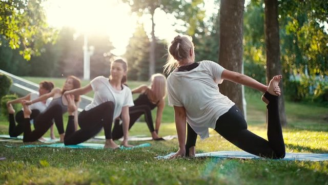 Group of women in sportswear are exercising in park moving from one position into another on yoga mats in slow motion. Group of people meditate in park on summer sunny morning under guidance of coach