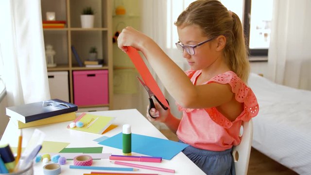childhood, creativity and hobby concept - creative girl cutting color paper with scissors at home