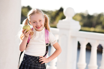 Laughing child girl 5-6 year old eating green fresh apple outdoors. Wearing school backpack and...