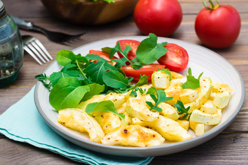 Slices of zucchini baked in spices with cheese on a plate with tomatoes and arugula on a wooden table