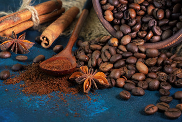 coffee grains scattered on a blue textural background, anise stars, cinnamon sticks and ground coffee in a wooden spoon.