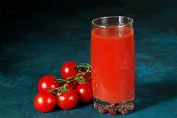 Glass cup with tomato juice and cherry tomatoes on a dark blue textural background.