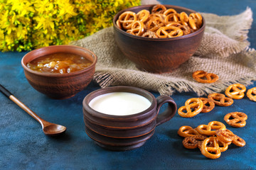 Still life in a rustic style. Crumbled pretzels on a textural background with milk in a clay cup, with apricot jam in a clay plate.