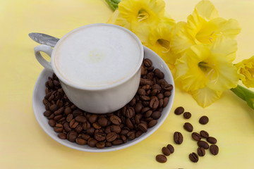 A cup of cappuccino coffee with milk crema and coffee beans on a yellow table, where there is a delicate yellow gladiolus flower. The concept of a soft and gentle summer cappuccino in yellow tones.