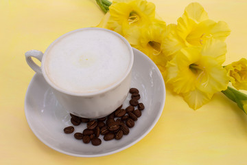 A cup of cappuccino coffee with milk crema and coffee beans on a yellow table, where there is a delicate yellow gladiolus flower. The concept of a soft and gentle summer cappuccino in yellow tones.