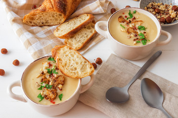 White wooden table with 2 bowls of cauliflower soup embellished with crushed hazelnuts, red pepper, smooth parsley herbs, Swiss twisted bread, frying pan with cauliflower, mortar and yellow flowers.