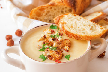 White wooden table with 2 bowls of cauliflower soup embellished with crushed hazelnuts, red pepper, smooth parsley herbs, Swiss twisted bread, frying pan with cauliflower, mortar and yellow flowers.