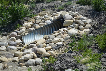 Metal drainage culvert underneath road surrounding by rocks.