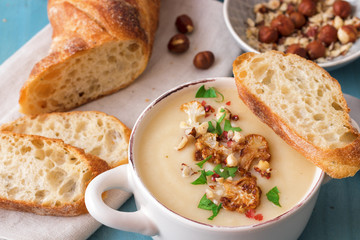 Blue wooden table with 2 bowls of cauliflower soup embellished with crushed hazelnuts, red pepper, smooth parsley herbs, Swiss twisted bread, frying pan with cauliflower, mortar and yellow flowers.