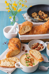 Blue wooden table with 2 bowls of cauliflower soup embellished with crushed hazelnuts, red pepper, smooth parsley herbs, Swiss twisted bread, frying pan with cauliflower, mortar and yellow flowers.