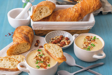 Blue wooden table with 2 bowls of cauliflower soup embellished with crushed hazelnuts, red pepper, smooth parsley herbs, Swiss twisted bread, frying pan with cauliflower, mortar and yellow flowers.