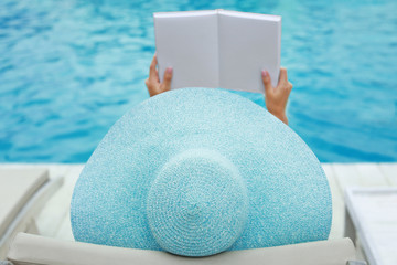Fototapeta na wymiar Beautiful young woman reading book while relaxing on sun lounger near swimming pool