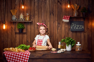 Smiling girl wearing white T-shirt with checkered apron and headband preparing dough with roller pin on table filled with ingredients for pizza, looking at camera in stylish wooden kitchen.