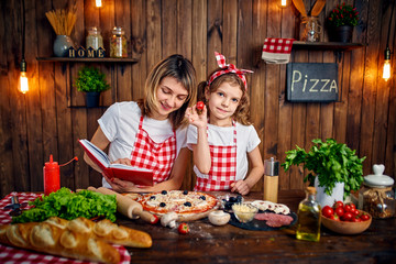 Happy mother and daughter wearing white T-shirts and checkered aprons cooking pizza together and decorating with mushrooms on table filled with ingredients, in stylish wooden kitchen.
