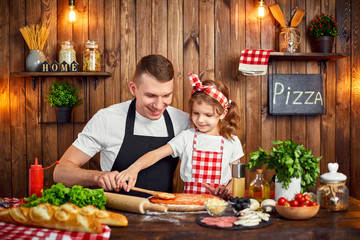 Happy father and daughter wearing aprons smiling and spreading sauce by dough with wooden spoon while cooking pizza in stylish kitchen interior