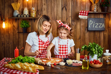 Happy mother, daughter wearing white T-shirts and checkered aprons cooking pizza together and decorating with mushrooms on table filled with ingredients in stylish wooden kitchen.