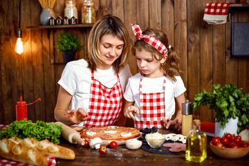 Happy mother and daughter wearing white T-shirts and checkered aprons cooking pizza together and decorating with mushrooms on table filled with ingredients in stylish wooden kitchen.