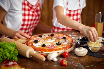 mother and daughter wearing white T-shirts and checkered aprons cooking pizza together and decorating with mushrooms on table filled with ingredients in stylish wooden kitchen