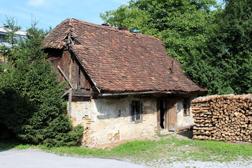 Fototapeta na wymiar Abandoned small family house with cracked walls and dilapidated facade covered with partially missing roof tiles surrounded with dense trees and prepared firewood in front on warm sunny spring day