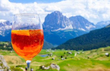 View of the traditional Italian alcoholic drink Aperol Spritz on the background of colorful Italian meadows and the Dolomites Alps mountains. village St. Cristina di Val Gardena Bolzano Seceda, Italy.