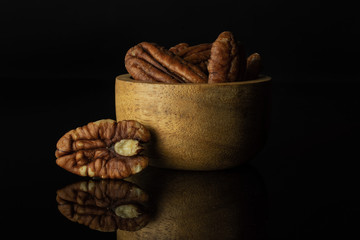 Lot of whole fresh brown pecan nut half with one near in a wooden bowl isolated on black glass