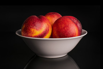 Group of three whole fresh red nectarine in a white ceramic bowl isolated on black glass