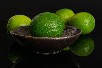 Group of four whole fresh green lime three are in the back and one is in the front in a dark ceramic bowl isolated on black glass