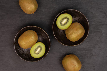 Group of four whole two halves of fresh green kiwifruit actinidia deliciosa placed symmetrically in a dark ceramic bowl flatlay on grey stone