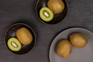Group of four whole two halves of fresh green kiwifruit actinidia deliciosa in a dark ceramic bowl on a gray ceramic plate flatlay on grey stone