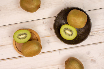 Group of four whole two halves of fresh green kiwifruit actinidia deliciosa on a round bamboo plate in a dark ceramic bowl flatlay on white wood