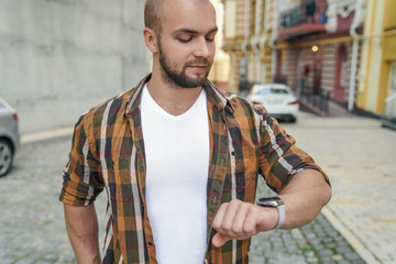 Obraz premium young good looking bold bearded guy standing outdoors near his house checking time making sure he is not late. smiling. wearing yellow shirt and jeans.